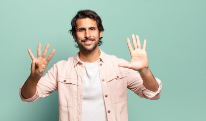 young handsome indian man smiling and looking friendly, showing number nine or ninth with hand...