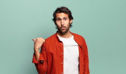 young handsome indian man looking astonished in disbelief, pointing at object on the side and...