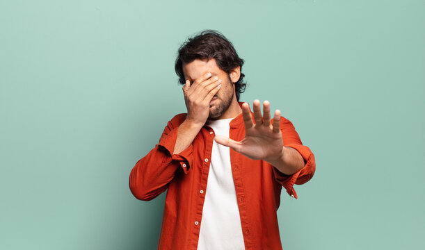 young handsome indian man covering face with hand and putting other hand up front to stop camera, refusing photos or pictures