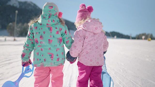 Slow motion - Two toddlers walking uphill with their shovel sledge and holding hands. Fun winter outdoor activity for kids. Right girl falls at the end of clip, oops 