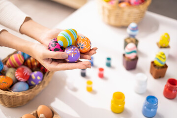 close up female hand holding easter egg with colorful patterns on the eggs in basket working on desk with brushes and paint cartilage, smiling joyfully having fun decoration celebrating easter holiday