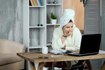 An emotional, relaxed young beautiful girl in a bathrobe and a towel on her head, sitting at a table watching movies or TV shows on a laptop and eating chocolate. Chill out and leisure concept.