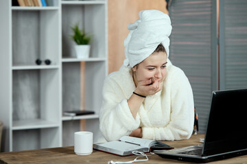 An emotional, relaxed young beautiful girl in a bathrobe and a towel on her head, sitting at a table watching movies or TV shows on a laptop and eating chocolate. Chill out and leisure concept.