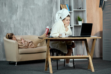 The concept of everyday student life. A girl sitting at a table in a white robe with a towel on her head and watching a movie or TV series on a laptop, and her friend is resting lying on the couch.