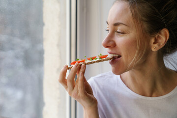 Happy woman eating rye crisp bread with creamy vegetarian cheese tofu, cherry tomato and rucola micro greens, sitting at home and looking at window. Healthy food, gluten free, diet concept.  - obrazy, fototapety, plakaty