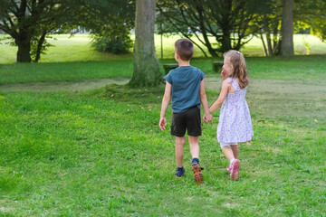 Fototapeta premium children walk in a summer park, a boy in shorts and a girl of 6-7 years old in dress are walking together on playground, holding hands, concept of active lifestyle, children's friendship