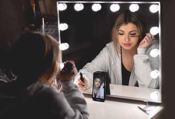 Young teenage girl applying mascara while live streaming in front of vanity mirror with lights