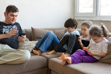 father and his children are sitting on the sofa. Playing internet games. they all have mobile phones and digital tablets in their hands