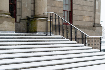 Large round concrete columns at the top of marble steps with black wrought iron rails to a legal building. The government building has a tall red door.  The steps are covered in fresh white snow. 