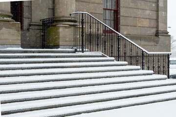 Large round concrete columns at the top of marble steps with black wrought iron rails to a legal building. The government building has a tall red door.  The steps are covered in fresh white snow. 