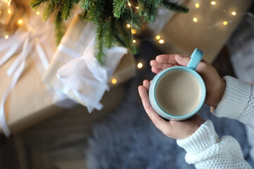 Woman holding cup of cocoa near Christmas tree indoors, top view