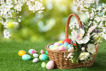 Wicker basket with Easter eggs and beautiful flowers on green grass outdoors, space for text