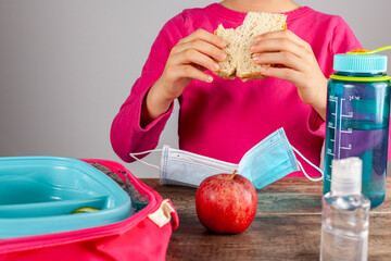 Closeup image with eating lunch at school concept during the phased reopening after COVID-19 pandemic closures. Girl with face mask removed eats sandwich snack with hand sanitizer on the table