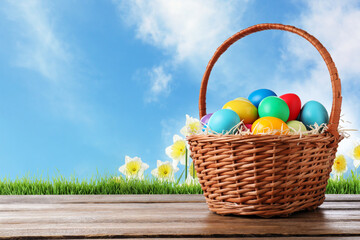 Colorful Easter eggs in wicker basket on wooden table outdoors, space for text