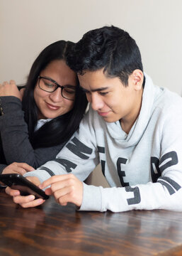 Young Mom With Latino Teenage Son Watching Cell Phone. Concept Children Teach Their Parents To Use Technology.