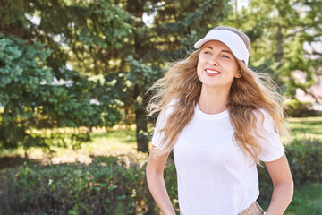 Pretty young woman wearing tennis hat. White t-shirt mockup. Female sport player. Female person posing outdoors. Happy smiling portrait. Nature green background. Long blond hair. Wind summer day