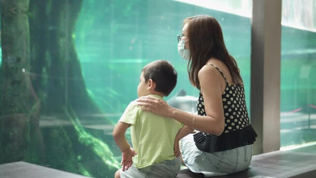 Mother and son at the science and natural musem looking at fish. Asian mom and child having educational family time. Young woman wearing face mask at a public place due to coronavirus outbreak
