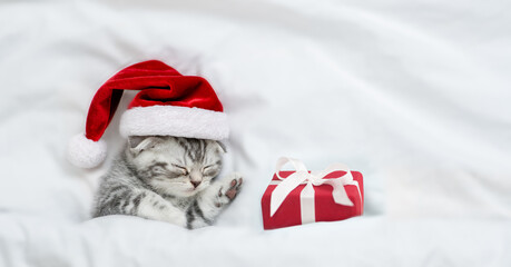 Cute kitten wearing red santa's hat sleeps with gift box under a white blanket on a bed. Top down view. Empty space for text