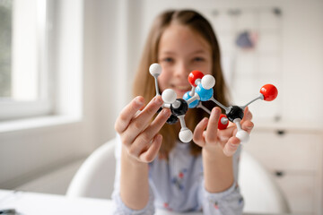 pretty blonde schoolgirl is learning chemistry with the help of a molecule model and is sitting at...