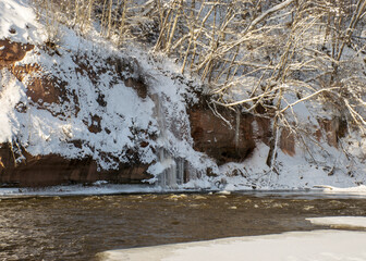 sunny day, landscape with red sandstone cliffs that are snowy with snow, frozen icicles on the cliff wall, icefall on the cliff wall, frozen river, Gauja, Kuku cliffs, Latvia