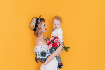 A woman in a dress holding a little boy in her arms and holding a beautiful bouquet of flowers in front of a yellow background. Holiday gift.