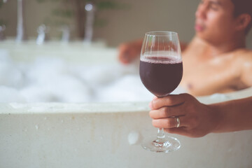 Close up a glass of wine in hand.  Asian man hold cold wineglass by hand in bathtub with soap bubbles. Selected focus. Holiday and relaxing time concept.