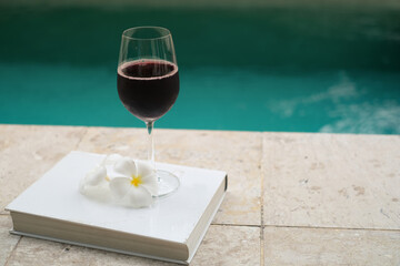 Glass of red wine and white frangipani tropical on a book near a swimming pool in morning of summer. Summer travel concept. Vacation and holiday concept. 
