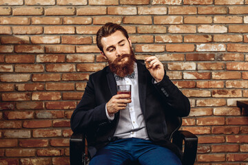 Portrait of a pretty amazing man with a mustache and a beard sitting on a chair and holding a glass...