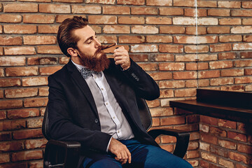 Portrait of a sharming brutal amazing man sitting on a chair and setting fire to a cigar against the background of a brick wall. Cigar concept