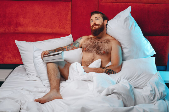 Amazing sexy muscular boy with a bushy beard and long hair and tattoos with a book in his hands smiling and sitting on a bed with gray linen and pillows against a black wall with a red velvet insert. 