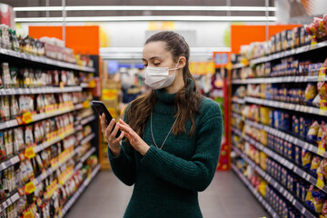 Woman wearing medical mask using smart phone while shopping in supermarket. Reading shopping list.