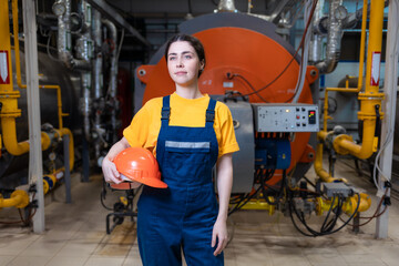 The concept of industrial manufactory. A young smiling woman in uniform holds a safety helmet in her hand and poses while standing in the boiler room