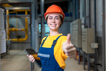 Industrial production. A female engineer in uniform and a protective helmet, holding a tablet in her hands, gives a thumbs up. The concept of equality and success