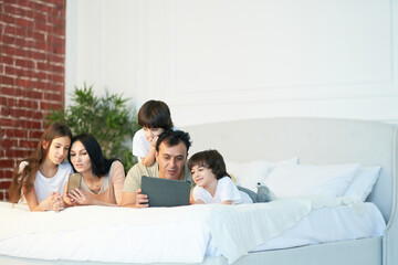 Technologies. Happy latin family with cute little kids using digital devices, lying on the bed together. Parents and children looking at the screen while having video call in the morning