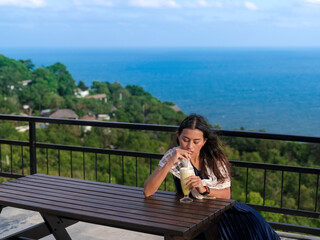 woman sit at wooden table drinking pineapple frappe on terrace with sea view on mountain peak at Koh Tao island, Suratthani, Thailand - Powered by Adobe
