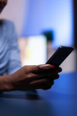 Close up of dark skinned woman chatting using smartphone in the evening from home office working remote. Employee using modern technology network wireless doing overtime.