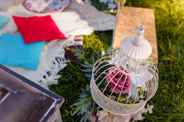 Soft focused shot of decorative bird cage filled with flowers, blurry cushions on plaid. Outdoors picnic party, birthday, proposal, anniversary.