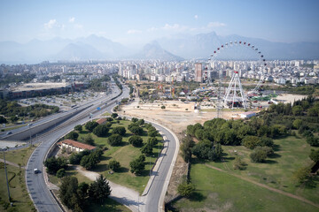 Fototapeta premium Urban landscape of Antalya town. Panoramic view of city buildings, road, Ferris wheel and mountains in the background.