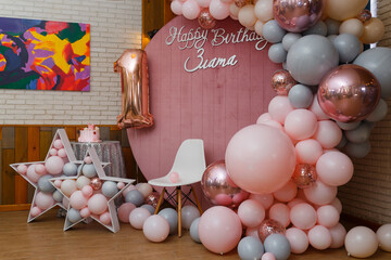 Beautiful festive decorations, pink and grey balloons arch, wooden stars, white chair and number one balloon on wooden round background. Little 1 year old girl birthday party photo zone.