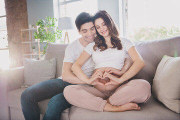 Photo of dreamy adorable good mood couple expecting child sitting couch embracing belly showing fingers heart inside indoors