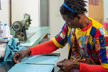 african tailor dressed in traditional clothes and fabrics at work in his workshop, production of...
