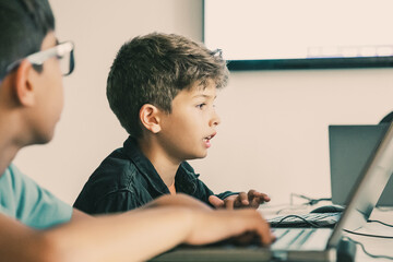 Caucasian boy reading task aloud during lesson. Classmates and pupils sitting at table in school and using laptops for study. Knowledge, childhood, communication and digital education concept
