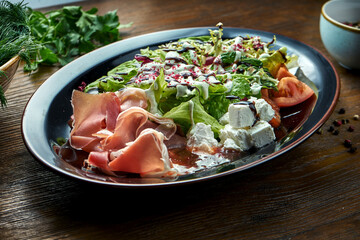 Delicious and dietary salad with jamon, feta cheese, tomatoes, served in a black plate on a wooden background. Restaurant food