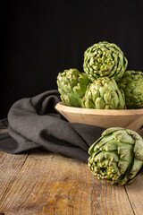 Top view of green artichokes in wooden bowl with black cloth, with selective focus, on wooden table, black background, vertical, with copy space