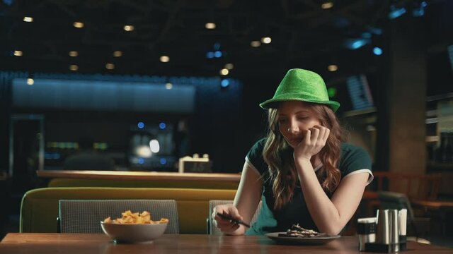 Pretty young woman waiting for someone, worried, looking at the phone and wearing an Irish green hat. Celebrating Saint Patrick's Day in a pub. 