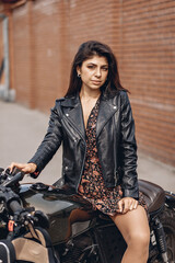 Fototapeta na wymiar Portrait of an amazing lovely adult female model wearing a black leather jacket and dress and sitting on a black motorcycle posing outdoors against a brick wall. Hobby ​​concept