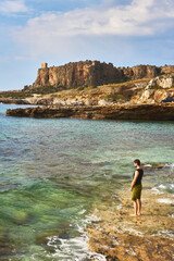 young caucasian man standing with feet int the mediterrenian sea near San vito lo Capo in Sicily looking toward the distance with ancient fortress in the background, 