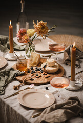 Romantic table setting, home lockdown party concept. Rose wine in bottle and glasses, cheese board, nuts, jam, dinnerware, cutlery, candles on light linen tablecloth, selective focus