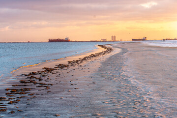 Ships at sea at sunset, viewed from a flat shoreline with ice on the sand with a high rise building in the background, Bolivar Flats, Texas