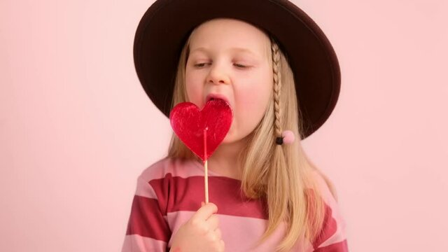 Little girl child enjoying eating licks sweet candy on a heart shaped lollipop stick. Selective focus.